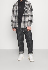 Schwarz-weiß karierte Flanellshirt, getragen über einem schwarzen Hoodie. Gestreifte schwarze Hose mit hochgekrempelten Säumen, kombiniert mit weißen Sneakern.