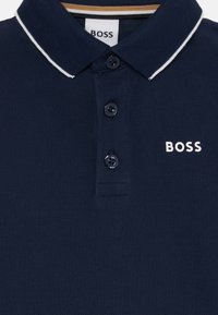 Tamsiai mėlyna polo marškinėlių su tekstūruotu audiniu. Yra trijų mygtukų apykaklė, balti akcentiniai juostelės ant apykaklės ir balta "BOSS" logotipas.
