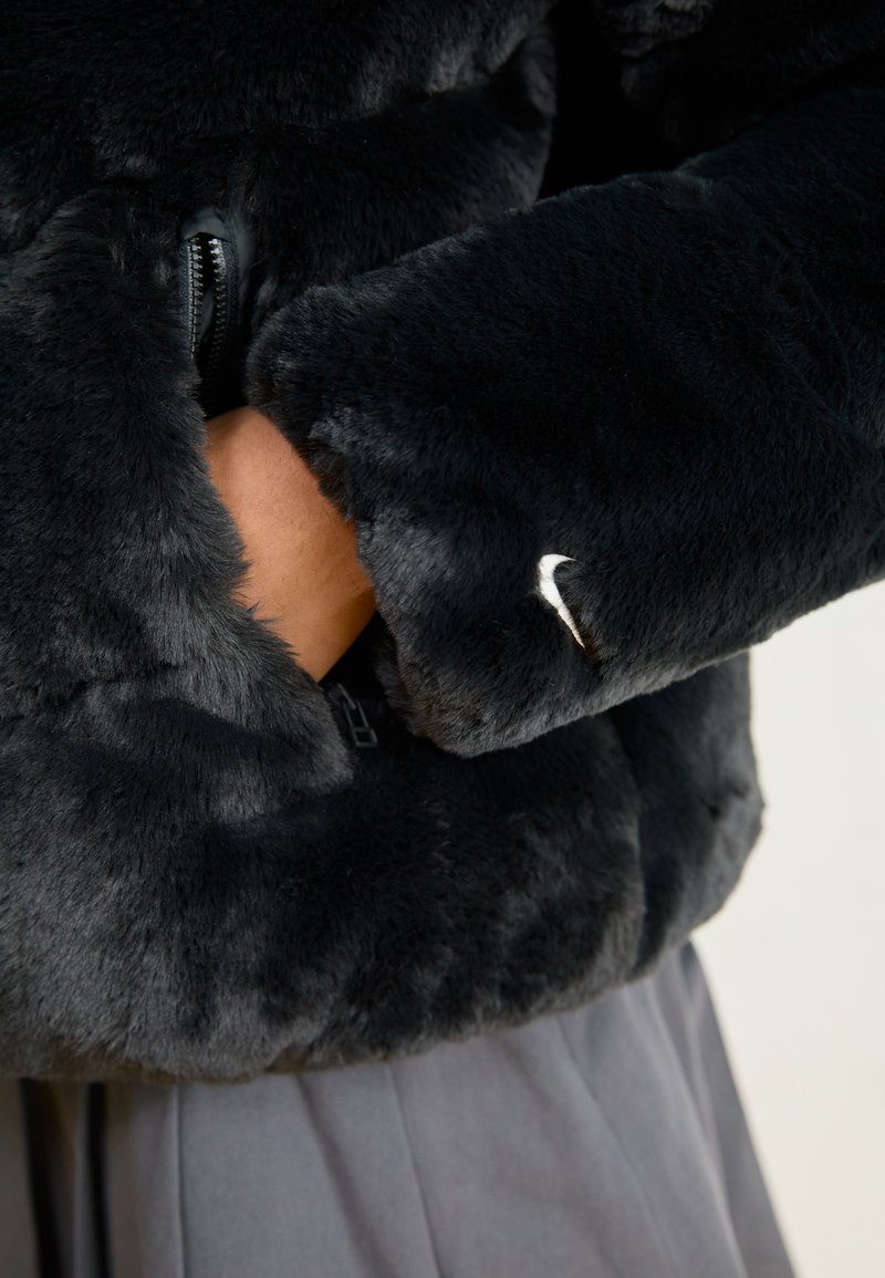 Veste en fausse fourrure noire avec une poche zippée et un logo Nike blanc sur la manche. Présente une texture douce et un design moelleux.