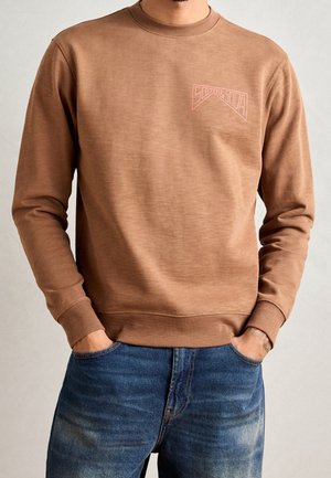 Mann trägt braune Langarm-Sweatshirt mit pinkem "Scotch & Soda"-Logo auf der Brust und blaue Jeans, Hände in den Taschen.