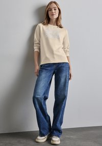 Světle béžová mikina s bílými grafickými detaily, kombinovaná s volnějšími modrými džínami a béžovými teniskami. Hladká textura, ležérní design.