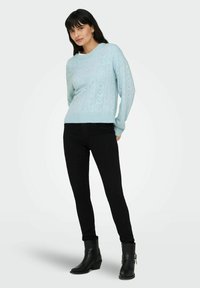 Pull en tricot bleu clair avec un motif texturé comprenant des rayures verticales et des formes de cœur, porté avec un jean skinny noir et des bottes noires.