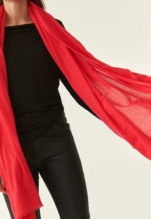 Femme portant un haut et un pantalon noirs avec une écharpe rouge vif flottant sur son bras tendu, sur un fond uni.