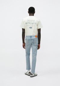 T-shirt color crema con scritta "DENIM ATTITUDE" in grigio, abbinata a jeans in denim azzurro chiaro. Imbottitura in pelle marrone chiaro sulla cintura. Calzature casual.