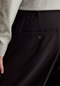 Černé kalhoty s jednoduchým designem, látkou s hladkým texturováním a zadní kapsou se zapínáním na knoflík. Viditelný elastický pas.