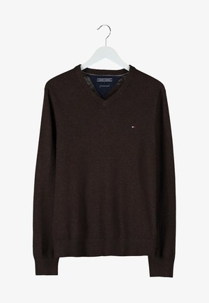 Trench beige sopra un maglione marrone scuro, abbinato a jeans neri e stivali marroni. Il cappotto presenta bottoni doppiopetto e colletto svasato.