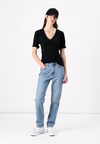 Sort kortærmet top med V-hals, parret med lyseblå jeans og hvide sneakers. Modellen har en sort kasket på og står foran en hvid baggrund.