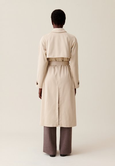 Cappotto trench beige con vita regolabile, design doppiopetto e spacco posteriore. Il modello indossa pantaloni a coste marrone chiaro.