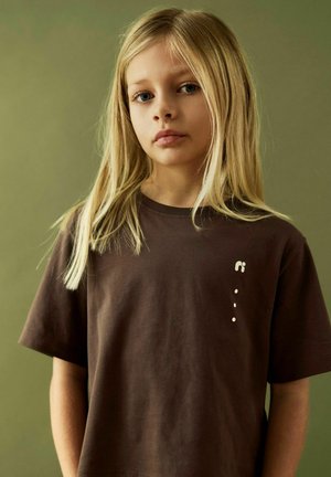 Blond child with blue eyes wearing a brown T-shirt with small white logo, standing against a plain green background, facing forward.
