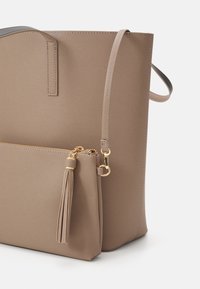 Sac à main beige texturé avec une pochette détachable assortie comprenant une fermeture éclair dorée et un détail pompon en cuir sur fond blanc.