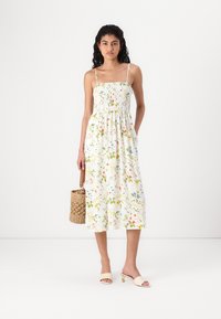 Λευκό φλοράλ φόρεμα με ρυθμιζόμενες τιράντες, smocked μπούστο και ρομαντική φούστα με επίπεδα; συνδυασμένο με μπεζ σανδάλια και μια υφαντή τσάντα.