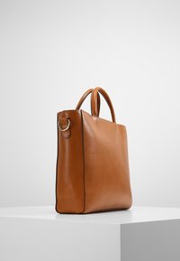 Sac fourre-tout en cuir marron, au toucher lisse, à la forme structurée et avec deux poignées. Présente un accent en métal argenté sur le côté.
