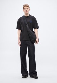 Μαύρο υπερμεγέθες μπλουζάκι με μπλε κεντημένο λογότυπο, συνδυασμένο με σκούρο μπλε τζιν παντελόνι φαρδιάς γραμμής και μαύρη τσάντα ώμου. Ομαλές υφές υφάσματος.