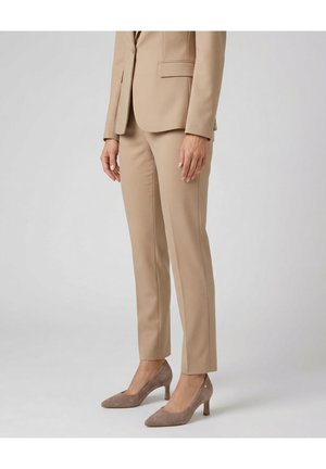 Vrouw die een beige op maat gemaakt pantalonpak draagt met bijpassende blazer en taupekleurige suède puntige hakken, staand tegen een effen lichtgrijze achtergrond.