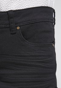 Jean en denim noir au design ajusté, avec une poche avant, des détails en métal doré et un tissu texturé avec des coutures visibles.