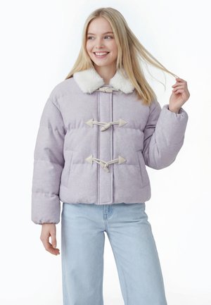 Ung kvinde smiler iført en lys lilla dunjakke med hvid pelskrave og knapper samt lyseblå jeans, holder en tot hår.