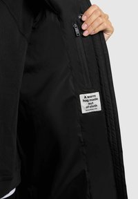 Interior de una chaqueta negra que muestra un bolsillo con cremallera y una etiqueta con texto. Material suave con un acabado mate, presenta costuras en contraste.