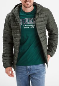 Chaqueta de plumas verde con diseño acolchado horizontal, cremallera delantera y capucha fija, llevada sobre una camiseta azul-verde y unos vaqueros.
