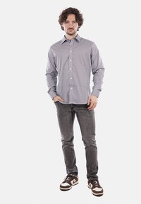 Camicia button-up con un motivo geometrico blu e bianco, maniche lunghe e una vestibilità sagomata, abbinata a jeans grigi e sneakers marroni.