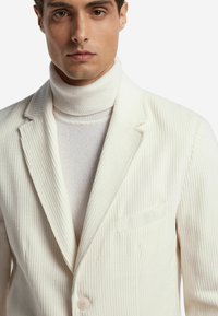 Weißes geripptes Blazer mit Revers, einem einzigen Knopfverschluss und einer linken Brusttasche, kombiniert mit einem hellen Rollkragenpullover.