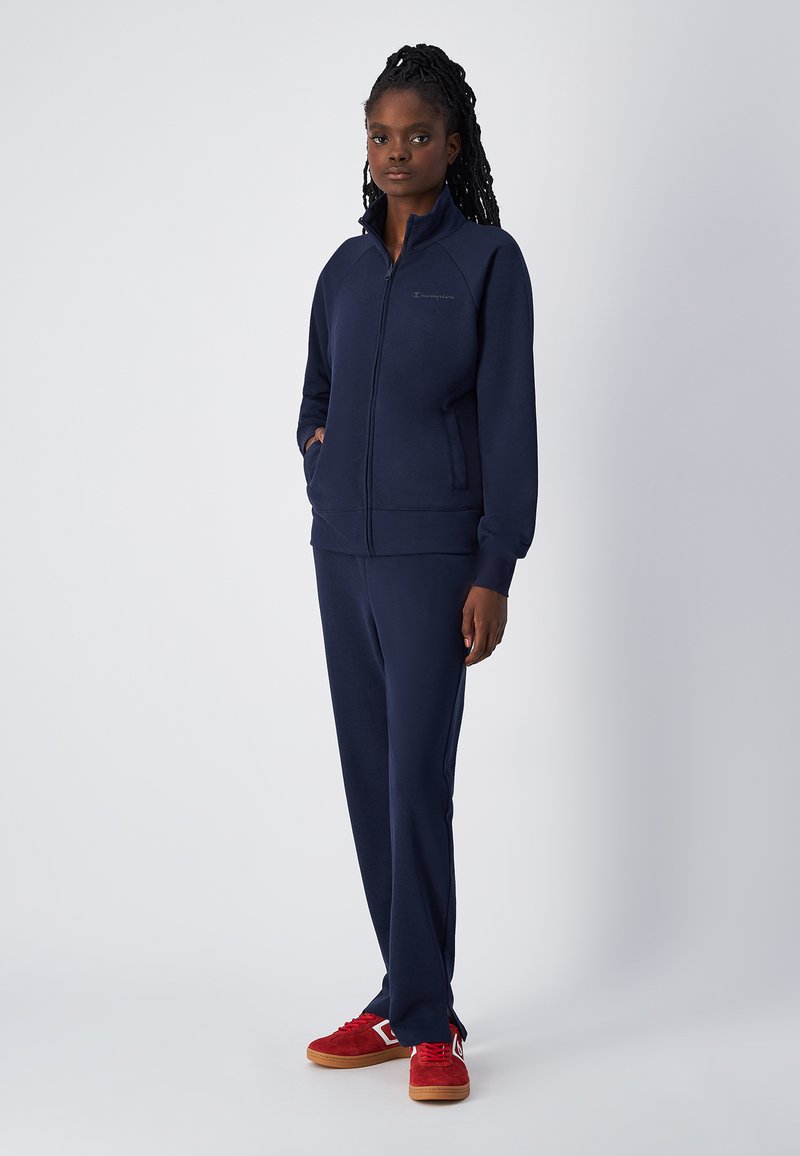 Marineblauw fleece trainingspak met een ritssluiting aan de voorkant, ribgebreide manchetten en tailleband, gecombineerd met rode sneakers met witte accenten en een rubberen zool.