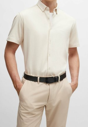 Homme portant une chemise crème à manches courtes boutonnée, rentrée dans un pantalon beige avec une ceinture noire, les mains dans les poches.