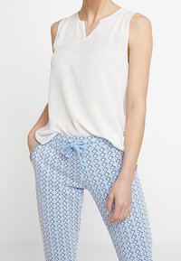 Blouse blanche sans manches avec col en V, associée à un pantalon bleu clair à motifs avec un design en losange et une taille à cordon.