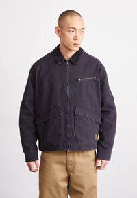 Dark navy jacket made of sturdy fabric with a zip front, two large front pockets, and a small chest pocket with zipper. Corduroy collar detail.