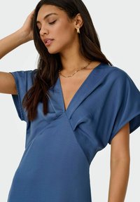 Niebieska sukienka krótki rękaw z miękkiej, gładkiej tkaniny; ma głęboki dekolt w kształcie litery V i plisowany przód. Noszona jest masywna złota naszyjnik.