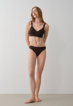 Une femme présente un soutien-gorge et une culotte noirs Calvin Klein, debout pieds nus devant un mur clair et uni.