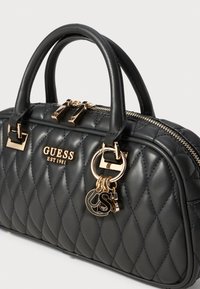 Black quilted handbag with gold hardware, dual top handles, zip closure, and logo detail. Features a decorative charm on the side.