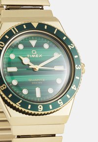 Montre-bracelet en quartz doré avec un cadran rayé vert, des index horaires blancs, des aiguilles dorées et une lunette verte, avec un bracelet métallique.
