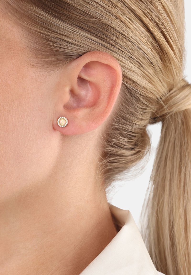 Ingrandimento dell'orecchio di una donna bionda che indossa un piccolo orecchino a bottone in opale circondato da piccole pietre trasparenti, con i capelli raccolti in una coda di cavallo.