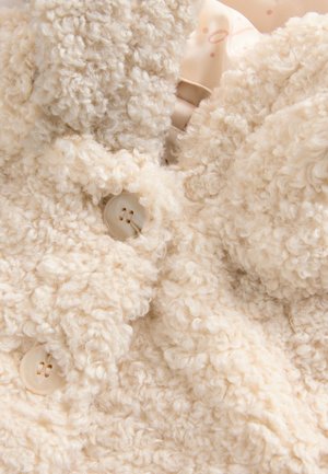 Palton bej crem din material pufos cu o textură moale. Are două nasturi deschiși la culoare și un design pufos, creț.