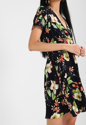 Woman wearing a black short-sleeve dress with colorful floral print and gold hoop earrings, hands clasped in front.