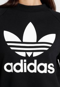 Czarna bluza z dużym białym logo Adidas w kształcie koniczyny oraz nazwą marki, z prążkowanym okrągłym dekoltem i miękką teksturą.