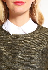 Kultakudottu villapaita, jossa on tekstuuripinta ja pyöreä kaulus, päällä valkoinen nappikauluspaita, jossa on klassinen kaulus.