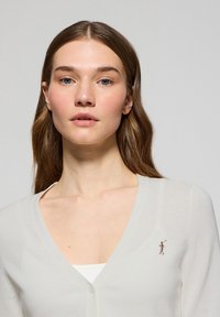 Cardigan de crema con escote en V y cierre de botones, que presenta una pequeña figura bordada en el lado izquierdo. Tejido suave y ligero.
