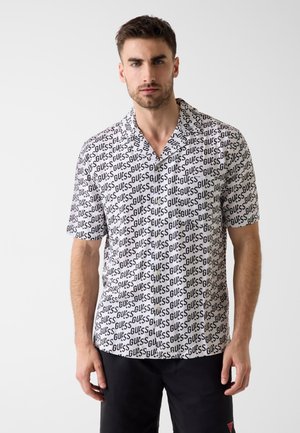 Homme portant une chemise blanche à manches courtes ornée du texte "GUESS" en noir répété, associée à un short noir, debout devant un fond uni.