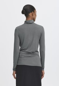 Donna con i capelli raccolti in un bun, che indossa un maglione grigio a maniche lunghe e una gonna nera, in piedi di spalle contro uno sfondo neutro.