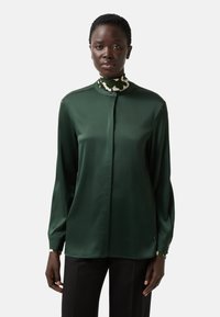 Dunkelgrüne Bluse mit langen Ärmeln, hohem Kragen und Knopfverschluss. Glatter Stoff mit dezentem Glanz. Musterakzent am Kragen.