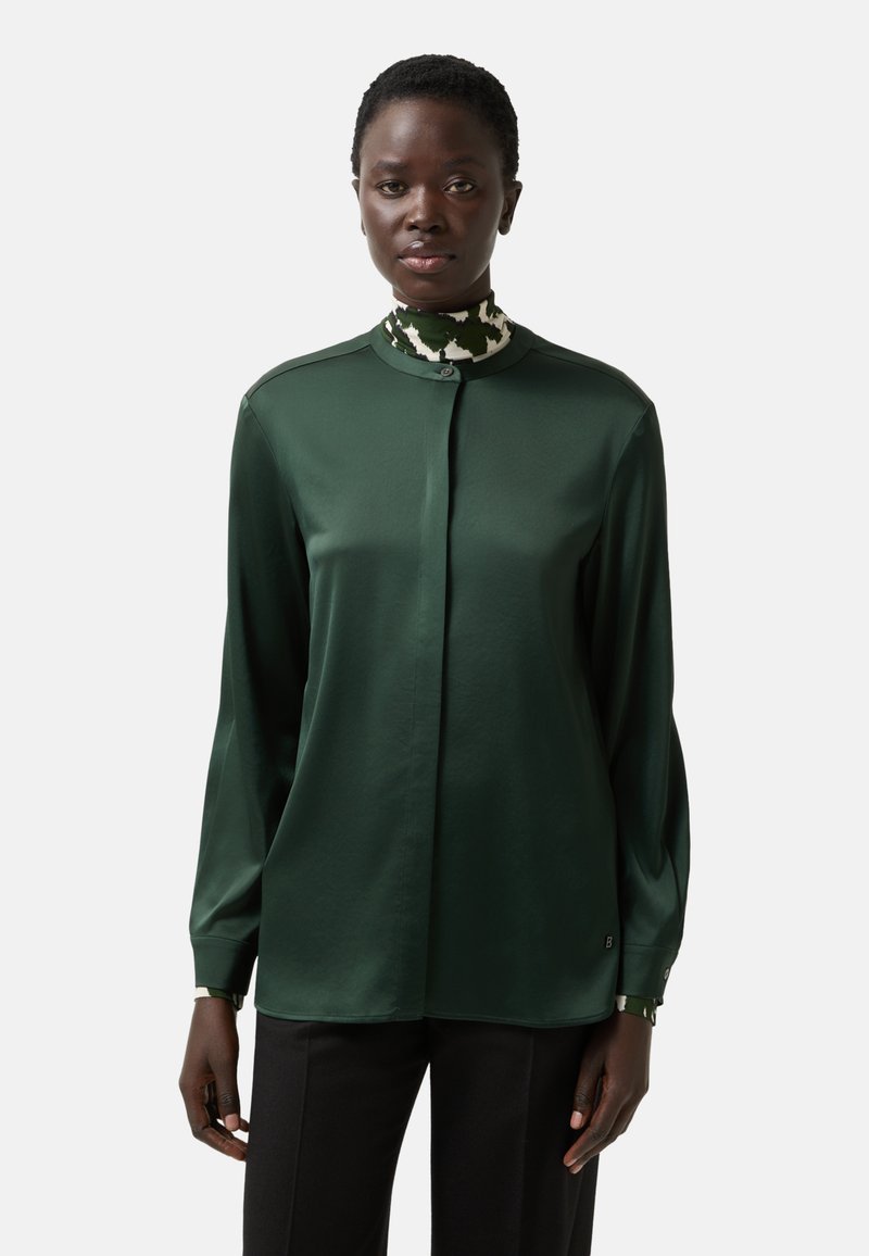 Dunkelgrüne Bluse mit langen Ärmeln, hohem Kragen und Knopfverschluss. Glatter Stoff mit dezentem Glanz. Musterakzent am Kragen.