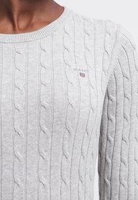 Pull gris en maille torsadée avec un col côtelé et un petit logo brodé sur la poitrine. Le matériau semble doux et texturé.