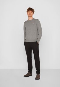 Grauer Strickpullover mit Rundhalsausschnitt, kombiniert mit schwarzen Hosen. Das Model steht mit den Händen in den Taschen und trägt braune Knöchelschäfte.