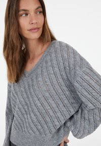 Grauer Strickpullover mit V-Ausschnitt, lockerer Passform und strukturiertem Muster mit horizontalen Streifen und kleinen offenen Webarten im Stoff.