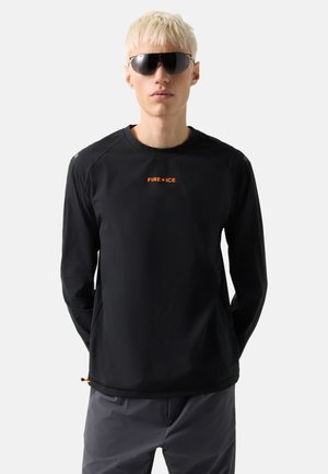 Ung mand med kort blondt hår iført sort langærmet trøje med orange "FIRE+ICE" tekst og mørke wraparound solbriller, hænderne bag ryggen.