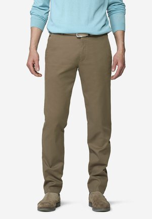 Mann trägt hellblauen Pullover, braune gerade geschnittene Hose mit Gürtel und passende braune Wildlederschuhe, steht vor weißem Hintergrund.