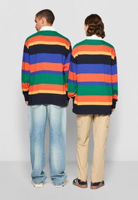 Stribede langærmede skjorter i rød, orange, blå, grøn og sort, båret med lyse jeans og khaki-bukser, vendt væk fra kameraet.