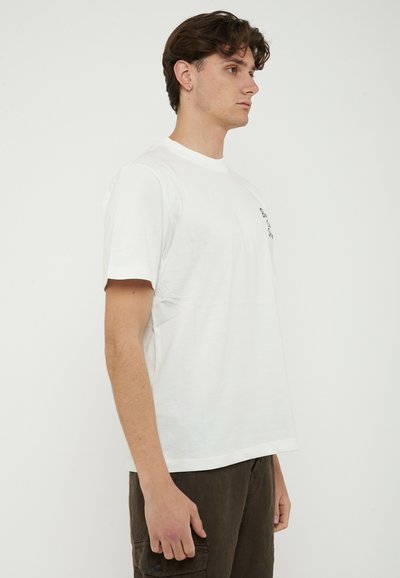 Giovane uomo con capelli castani corti in piedi di lato, indossa una semplice maglietta bianca e pantaloni scuri su uno sfondo bianco uniforme.