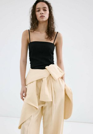 Woman with curly hair wearing a black sleeveless top and beige wide-leg pants with a matching jacket tied at the waist.